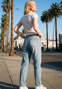 Joven mujer con camiseta blanca y vaqueros azul claro de pie en una calle soleada bordeada de altas palmeras, mirando hacia un lado.