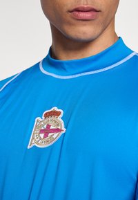 Modré sportovní tričko s vysokým límcem, zdobené bílým prošíváním a vyšívaným logem Real Club Deportivo La Coruña.