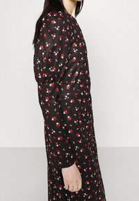 Kvinna iklädd en svart klänning med små röda och vita blommor, långa puffärmar, sedd från sidan mot en enfärgad bakgrund.