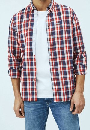 Homme portant une chemise à carreaux rouge, blanche et bleue déboutonnée sur un t-shirt blanc et un jean bleu, les bras détendus le long du corps.