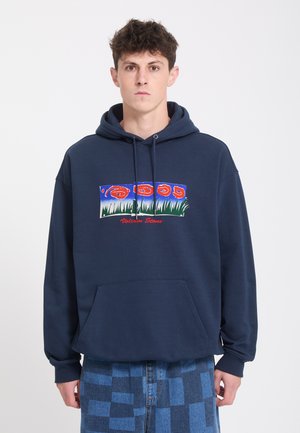 Jeune homme portant un sweat à capuche bleu marine avec un motif rouge et vert au-dessus du texte et un pantalon à carreaux bleu, debout devant un fond blanc uni.