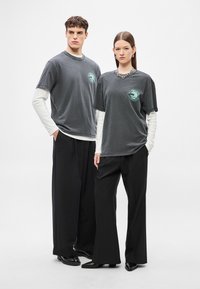 Αμφίεση unisex, γκρι βαμβακερά μπλουζάκια με εκτυπωμένο λογότυπο, φορεμένα πάνω από λευκά μακρυμάνικα μπλουζάκια. Συνδυασμένα με άνετα μαύρα παντελόνια και κομψά παπούτσια.