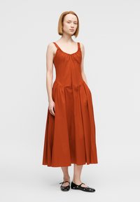 Φόρεμα μακρύ χωρίς μανίκια σε χρώμα σκουριάς, με σούρες στη λαιμόκοψη και στη μέση. Φαρδιά φούστα, κατασκευασμένη από ελαφρύ ύφασμα, με λεία υφή.