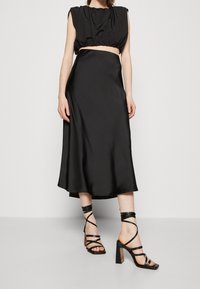 Robe en satin noir avec un haut sans manches froncé, une jupe mi-longue et des talons noirs à lanières avec un talon épais et un détail de maintien à la cheville.