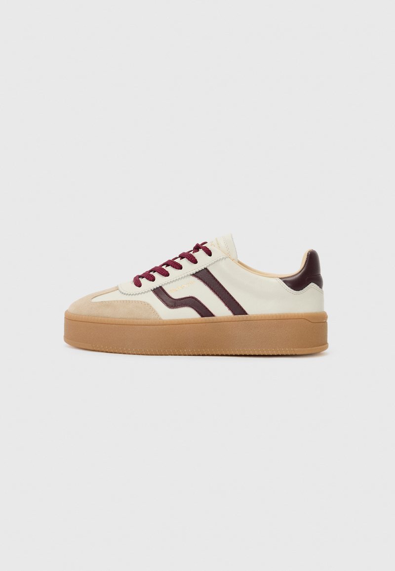 Sneaker med en kremfarget overdel, maroon detaljer, teksturert beige semsket skinn, og en tykk gummisåle. Har maroon snøringer og to parallelle striper.