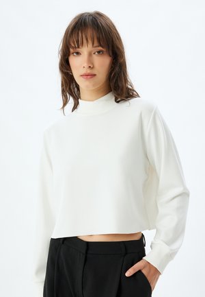 White cropped sweatshirt with a mock neck and ribbed cuffs. Soft texture, minimal design, and paired with black trousers.
