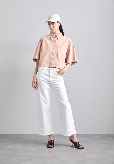 Lichtroze cropped overhemd met knopen en een zak, gecombineerd met witte wijdgevallen jeans en bruine open-teen hakken. Een plain witte pet maakt de look af.