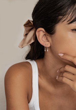 Orecchini a cerchio in argento con piccole perline pendenti, indossati da una persona dai capelli scuri, acconciati con un fiocco in satin beige. Lo sfondo è neutro.