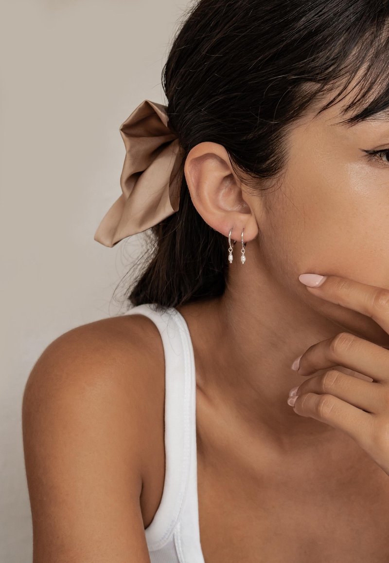 Boucles d'oreilles en argent en forme de hoop avec de petites perles pendantes, portées par une personne aux cheveux foncés, coiffés d'un nœud en satin beige. L'arrière-plan est neutre.