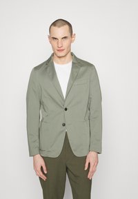 Helles grünes, aus Baumwollmischung gefertigtes Blazer mit slim Fit, zwei Fronttaschen, Reverskragen und einem einzelnen Knopfverschluss. Kombiniert mit einem weißen T-Shirt.