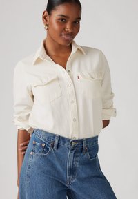 Camisa blanca de botones con cuello, dos bolsillos en el pecho, mangas arremangadas, combinada con unos vaqueros de cintura alta azules. Diseño minimalista y textura suave.