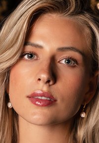 Close-up of a woman's face with light skin, long blonde hair, wearing gold hoop earrings with pearl accents. Glossy lips and clear skin.