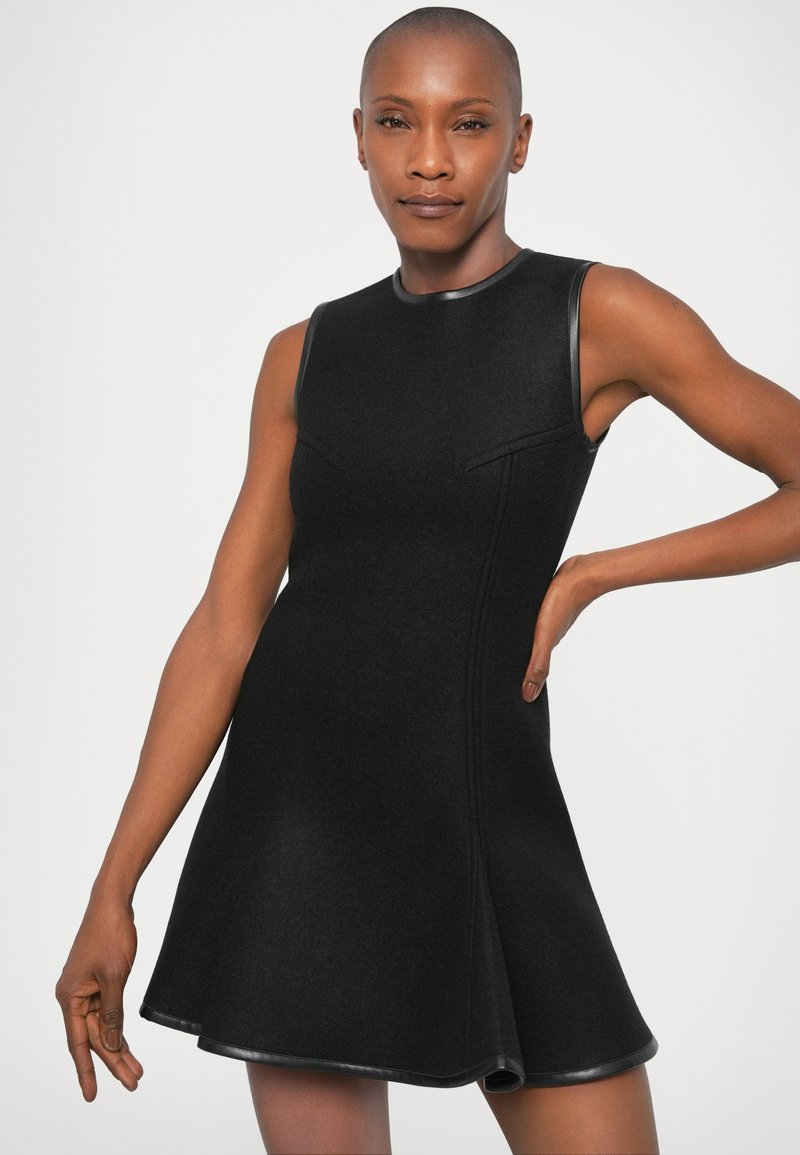 Robe noire sans manches avec une silhouette évasée, faite de tissu texturé, présentant un col en cuir lisse et des accents sur l'ourlet.
