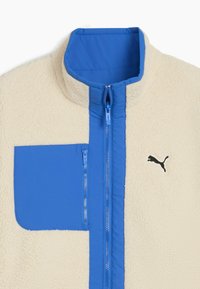 Kerma fleece takki, jossa on sininen nylon etupaneeli, vetoketjullinen tasku ja musta logo. Ominaisuuksiin kuuluu korkea kaulus ja kestävä rakenne.