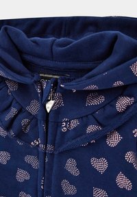 Námornícka mikina na zips s kapucňou, s ružovými srdiečkovými vzormi a volánovým golierom. Vyrobená z mäkkej, textúrovanej látky.