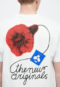 Biele tričko s veľkým červeným kvetinovým motívom a modrou pečiatkou s ilustráciou včely. Text je čiernou farbou a znie „thenew originals“.