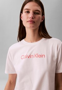 Femme aux cheveux bruns portant un t-shirt blanc avec "Calvin Klein" en texte rose sur la poitrine, debout devant un fond gris uni.