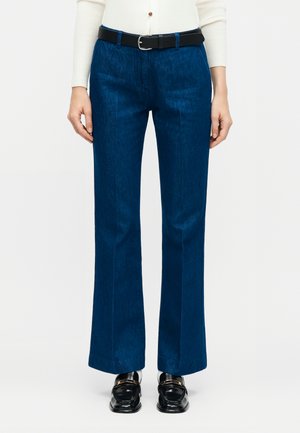 Kvinde iført mørkeblå jeans med lige ben, sort bælte, hvid ribbet bluse med lange ærmer og sorte loafers med gulddetaljer.