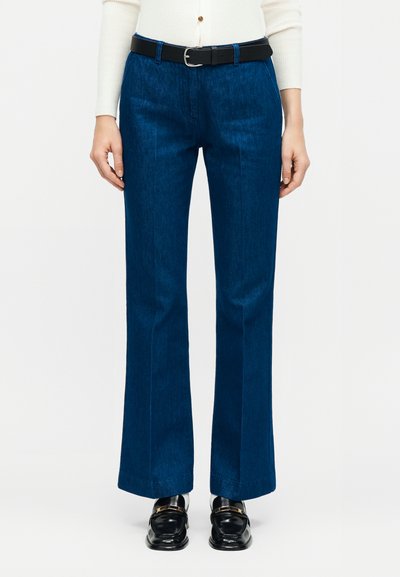 Femme portant un jean droit bleu foncé, une ceinture noire, un haut blanc à manches longues côtelé et des mocassins noirs avec ferrures dorées.