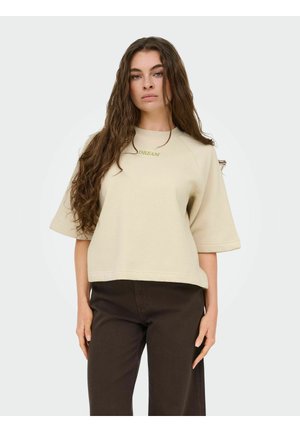 Mujer joven con cabello largo y ondulado, viste una camiseta beige de manga corta con la palabra "SUEÑO" y pantalones marrón oscuro, de pie frente a un fondo liso.