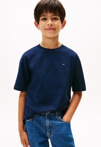 Boy with short dark hair wears navy blue short-sleeve T-shirt and blue jeans, standing with one hand in pocket against white background.