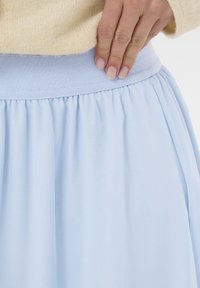 Světlomodrá sukně s nařaseným designem a širokým, texturovaným pasem v doplňujícím odstínu. Detailní záběr ukazuje texturu látky.