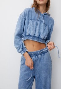 Sudadera de mezclilla azul recortada con cintura elástica y cordones, que presenta un bolsillo frontal. Combinada con joggers de mezclilla a juego.