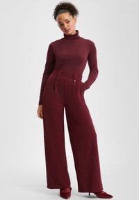 Burgunderfarbener Rollkragenpullover kombiniert mit hoch taillierten weit geschnittenen Hosen, die seitliche Taschen und zwei auffällige Vorderknöpfe aufweisen. Glatte Textur.