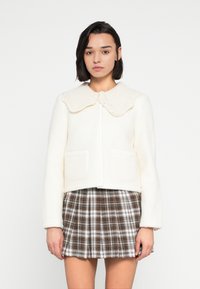 Λευκό σακάκι από μείγμα μαλλιού με ριμπ πλεκτό, κυματιστό γιακά και δύο μπροστινές τσέπες, σε συνδυασμό με καφέ καρό πλισέ φούστα.