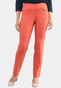 Pantalons couleur corail à coupe slim, dotés de deux poches zippées à l'avant et d'une texture lisse, assortis à des chaussures de couleur claire.