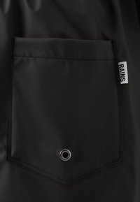 Il tessuto impermeabile nero presenta una tasca a forma di diamante con una superficie liscia, un occhiello argento e un'etichetta con "RAINS" sul lato.