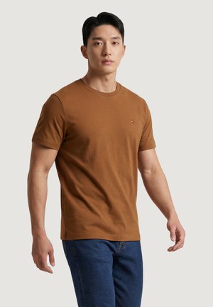 Uomo con t-shirt marrone a maniche corte e jeans blu, in piedi davanti a uno sfondo chiaro e uniforme, rivolto in avanti.