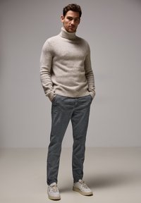 Světle šedý rolák z měkkého úpletu v kombinaci s tmavě šedými kalhotami a bílými teniskami. Modelka stojí s rukama v kapsách.