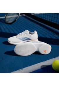 Λευκά παπούτσια τένις με ανοιχτομπλε ρίγες, υφή στη σόλα και πορτοκαλί λεπτομέρειες, τοποθετημένα σε μπλε γήπεδο κοντά σε μπάλα και ρακέτα τένις.