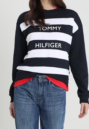 Frau trägt einen marineblau-weiß gestreiften Pullover mit dem Schriftzug "Tommy Hilfiger", darunter ein rotes Hemd und blaue Jeans.
