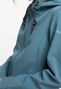 Veste zippée sarcelle avec capuche, présentant une texture lisse, des cordons de serrage ajustables et un logo sur le bras supérieur.