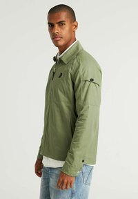 Groene jacket met een ritssluitingen aan de voorkant, twee borstzakken en met knopen sluitingen aan de mouwen. Gemaakt van een glad, duurzaam materiaal. Draag het over een wit shirt.