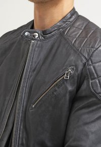 Schwarze Lederjacke mit gesteppte Schulterdetails, zippered Brusttasche und Druckknopfkragen, getragen von einer Person.