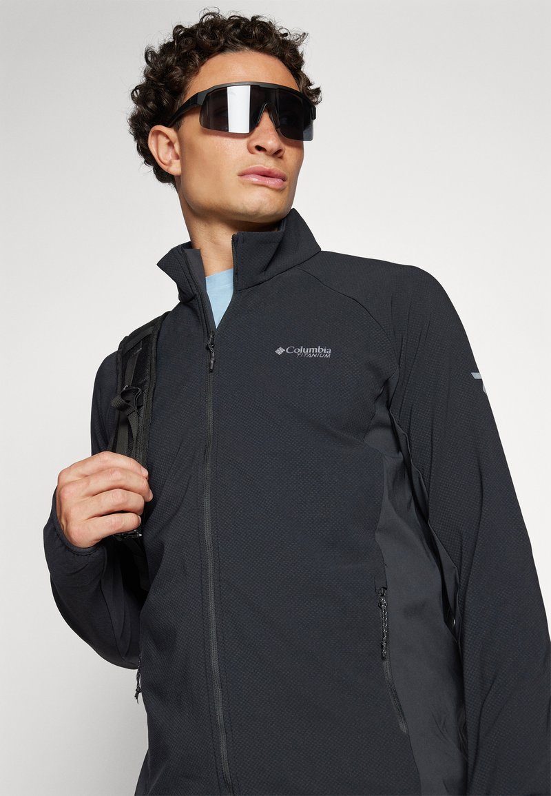 Schwarze Zip-Jacke aus strukturiertem Material, Columbia-Logo auf der Brust und mit einem eleganten Design. Das Modell trägt oversized schwarze Sonnenbrillen und hat einen Rucksack dabei.