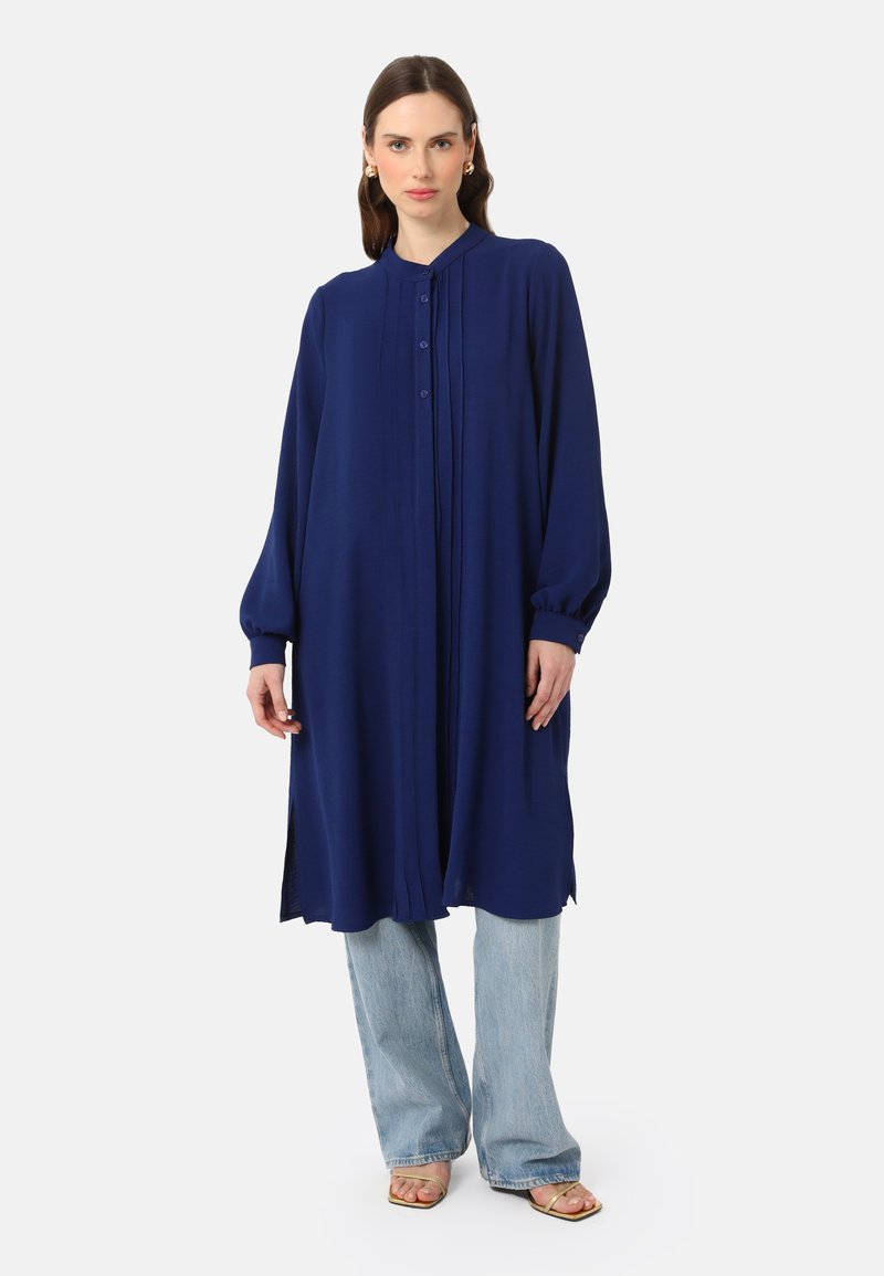 Abito lungo blu navy con colletto, bottoni a placket e dettagli plissettati; abbinato a jeans azzurri e sandali color nudo.