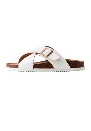 Sandalia blanca tipo diapositiva con suela de corcho marrón, tiras anchas cruzadas y una hebilla dorada sobre un fondo blanco.