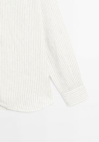 Chemise blanche à manches longues avec de fines rayures verticales noires et un poignet à rayures verticales sur un fond blanc uni.