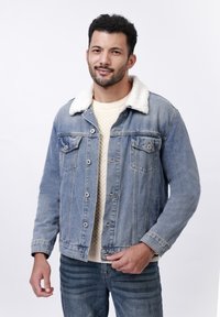 Lichtblauwe denim jas met een nepbont kraag, knoopsluiting en twee borstzakken. Draagt over een crèmekleurige trui.