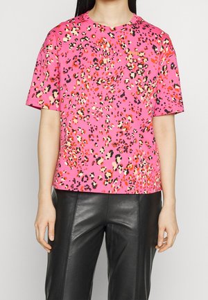 Vrouw draagt een felroze panterprint T-shirt met korte mouwen gecombineerd met zwarte leren broek, staand tegen een effen achtergrond.