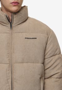 Šviesiai rudos kordūro vestuvinės striukės su paminkštintomis dalimis, aukštu apykakle ir juodu "PEGADOR" logotipu ant krūtinės; turi užtrauktuką.