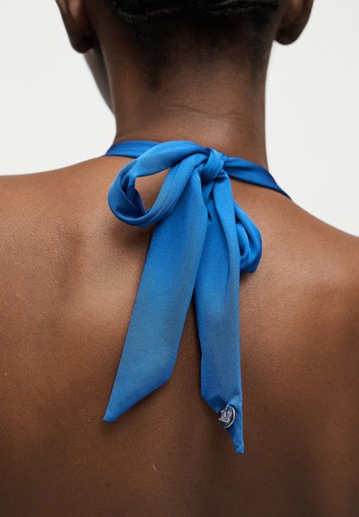 Μπλε ύφασμα δεμένο σε φιόγκο στο πίσω μέρος του λαιμού ενός ατόμου, με ένα μικρό στρογγυλό ασημένιο φυλαχτό προσαρτημένο σε ένα από τα άκρα του φιόγκου.