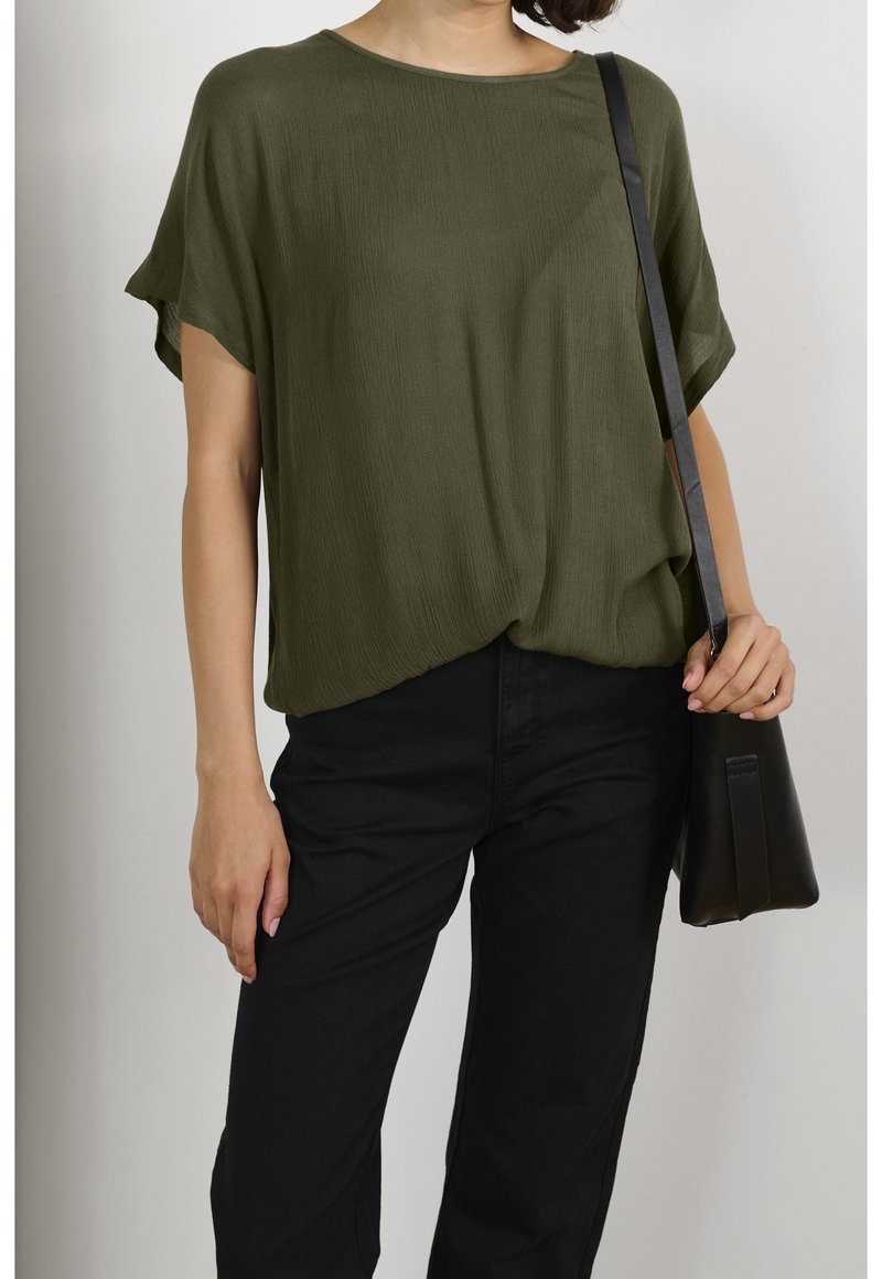 Top di un verde oliva, morbido e dalla vestibilità ampia, realizzato in un tessuto texturizzato. Abbinato a pantaloni neri e a una piccola borsa a tracolla nera.