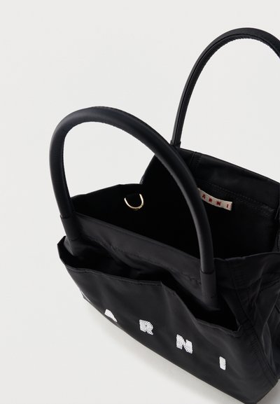Μαύρη υφασμάτινη τσάντα tote με καμπυλωτές λαβές και λευκόι παγιέτες μερικό κείμενο λογότυπου "ARNI" στο μπροστινό μέρος, εμφανίζεται ανοιχτή από το επάνω μέρος.