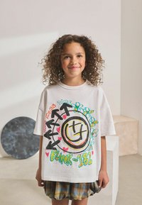 Fille aux cheveux bouclés portant un t-shirt graphique oversize blanc de Blink-182 et une jupe à carreaux, se tenant dans une pièce beige minimaliste.