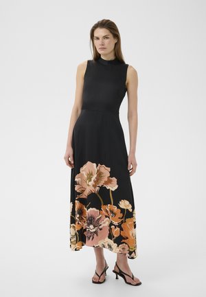 Vrouw in een mouwloze zwarte jurk met grote roze en oranje bloemenprint op de rok, gecombineerd met zwarte sandalen met hak, staand.